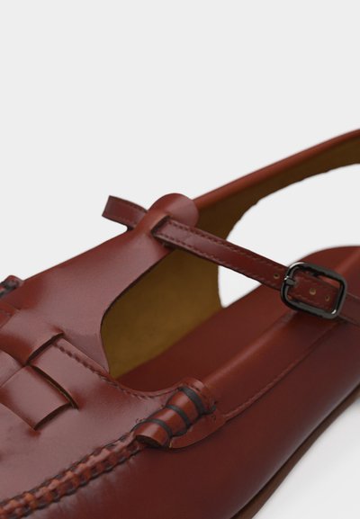 Καφέ δερμάτινο επίπεδο παπούτσι με T-λαιμό και κλείσιμο με πόρπη, με λεπτομερείς ραφές και υφασμένα δερμάτινα στοιχεία σε λευκό φόντο.