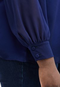 Μανσέτα μπλουζάκι σε σκούρο μπλε με μακριά μανίκια και δύο καλυμμένα κουμπιά σε άτομο που φορά σκούρο μπλε τζιν.