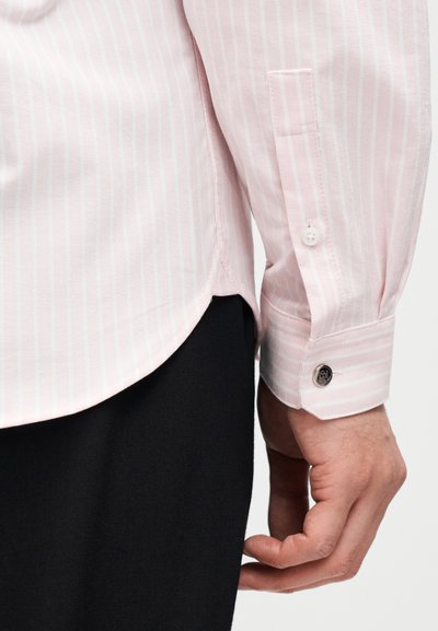 Ροζ ριγέ πουκάμισο με λευκές κάθετες ρίγες, μανσέτα με κουμπιά και ανοίγματα στις πλευρές. Το ύφασμα φαίνετ αι υφή με ομαλές λεπτομέρειες.