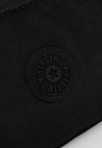 Zwarte stoffen tas met een ronde, gestructureerde zwarte rubberen logo met "KIPLING" en een sterontwerp in het midden. Gladde oppervlakte met stiksels.
