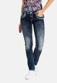 Schmal geschnittene, dunkelblaue Jeans mit einem ausgebleichten Finish, kontrastierenden Nähten und dekorativen Fronttaschen. Kombiniert mit weißen Sneakers.