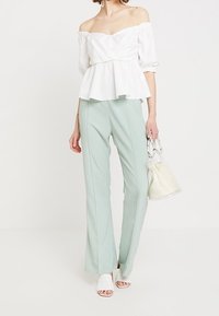 Bílý top s odhalenými rameny a peplum střihem se krátkými rukávy, ve spojení se světle zelenými elegantními kalhotami a bílými sandály na podpatku, držící průhlednou tašku.