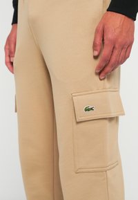 Beige kalhoty s boční kapsou na klopu, která nese zelené logo krokodýla, zobrazené s rukou osoby, která odpočívá blízko kapsy.