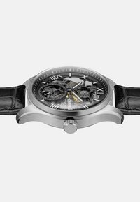 Mechanisch horloge met een roestvrijstalen kast, zwart skeleton wijzerplaat, Romeinse cijfers en een gestructureerde zwarte leren band. Heeft een kroon detail.