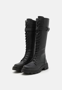 Bottes noires en cuir montant jusqu'aux genoux avec un laçage à l'avant, une semelle plate et des sangles décoratives. Présente une finition texturée et une couture robuste.