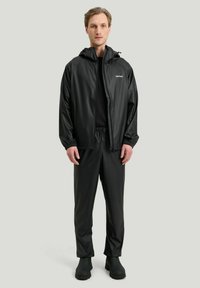 Svart vattentät jacka med huva, elastiska ärmslut och matchande byxor. Slät yta, minimal branding på jackan och gummistövlar.