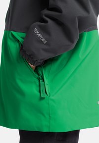 Bras dans une veste noire et verte avec un poignet élastique montrant la main à l'intérieur d'une poche zippée verte, manche étiquetée TEXAPORE.