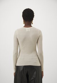 Beige langermet topp laget av et mykt, lett stoff med en tettsittende design og subtil logo på den øvre ryggen. Kombinert med svarte lærbukser.