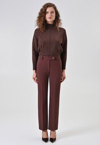 Maglione a collo alto marrone glitterato abbinato a pantaloni bordeaux a vita alta con dettaglio di bottoni laterali. Modella che indossa tacchi neri a punta.