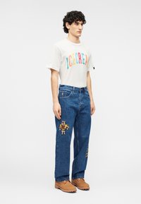 Λευκό μπλουζάκι με πολύχρωμο κείμενο "ICECREAM", συνδυασμένο με μπλε τζιν παντελόνι με κεντήματα αρκούδας και αστεριών, και καφέ μπότες.