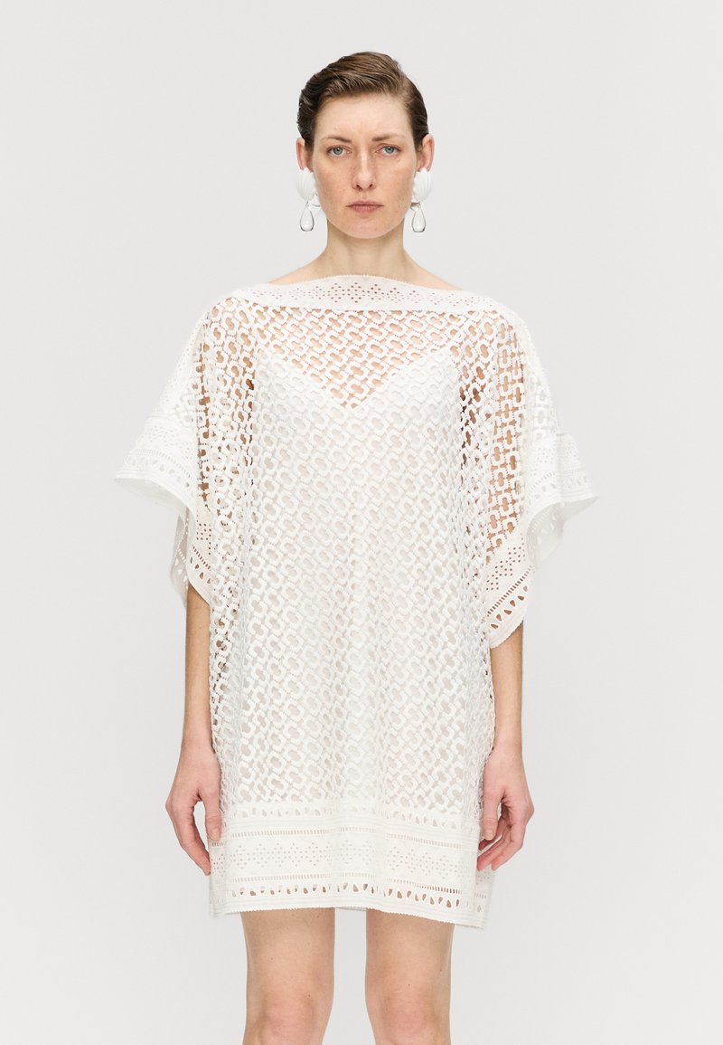 Rochie albă din dantelă, cu un croi lejer, având un model geometric, mâneci largi și o margine fantezistă. Detalii decorative includ decupaje.