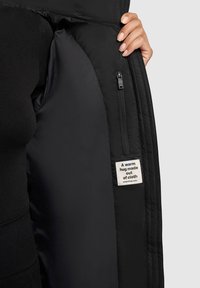 Zwart pufferjack binnenkant met een gladde textuur, een ritszak en een label met de tekst "Een warme omhelzing gemaakt van stof."
