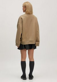 Beige oversized sweatshirt met gezakt schouders, gecombineerd met een zwarte, korte geplooide rok. Het model draagt kniehoge grijze sokken en zwarte schoenen.