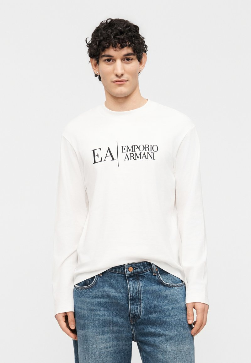 Νέος άνδρας με σγουρά μαλλιά που φοράει λευκό πο υκάμισο μακρυμάνικο Emporio Armani και μπλε τζιν, στέκεται μπροστά σε λευκό απλό φόντο.