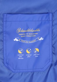 Kék vízálló szövetfolt arany szöveggel és ikonokkal, amelyek a vízállóságot, szélállóságot és a teljesen lezárt varrásokat jelzik.