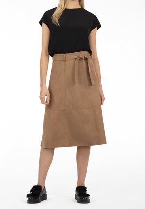 Donna che indossa una maglietta nera a maniche corte, gonna a linea A in camoscio color sabbia con cintura e mocassini neri con dettagli in pelliccia, in piedi su sfondo bianco.