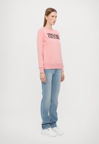 Rozā sporta krekls ar melnu "VERSACE JEANS COUTURE" logotipu, ribotām piedurknēm un apakšmalu; kopā ar gaiši ziliem džinsu biksēm un baltām čībām.