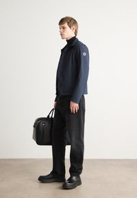 Mornarska zip-up jakna z okroglo logo nalepko, v kombinaciji s črnim пуловерjem, črnimi hlačami in črnimi čevlji na zavezovanje. Nosijo črno torbo.