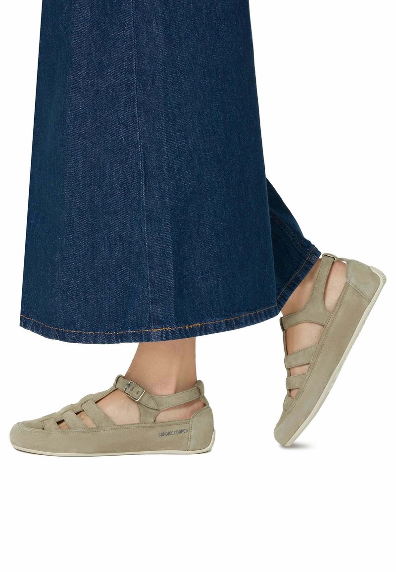 Chaussures en daim beige avec un design ouvert et une fermeture à sangle, associées à une longue jupe en denim foncé. Les chaussures présentent des accents texturés et une semelle plate.