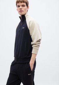 Σκούρο μπλε και εκρού μπουφάν με φερμουάρ, ελαστικά μανσέτες και γιακά, με λογότυπο στο στήθος, σετ με αντίστοιχο σκούρο μπλε παντελόνι φόρμας με τσέπη.