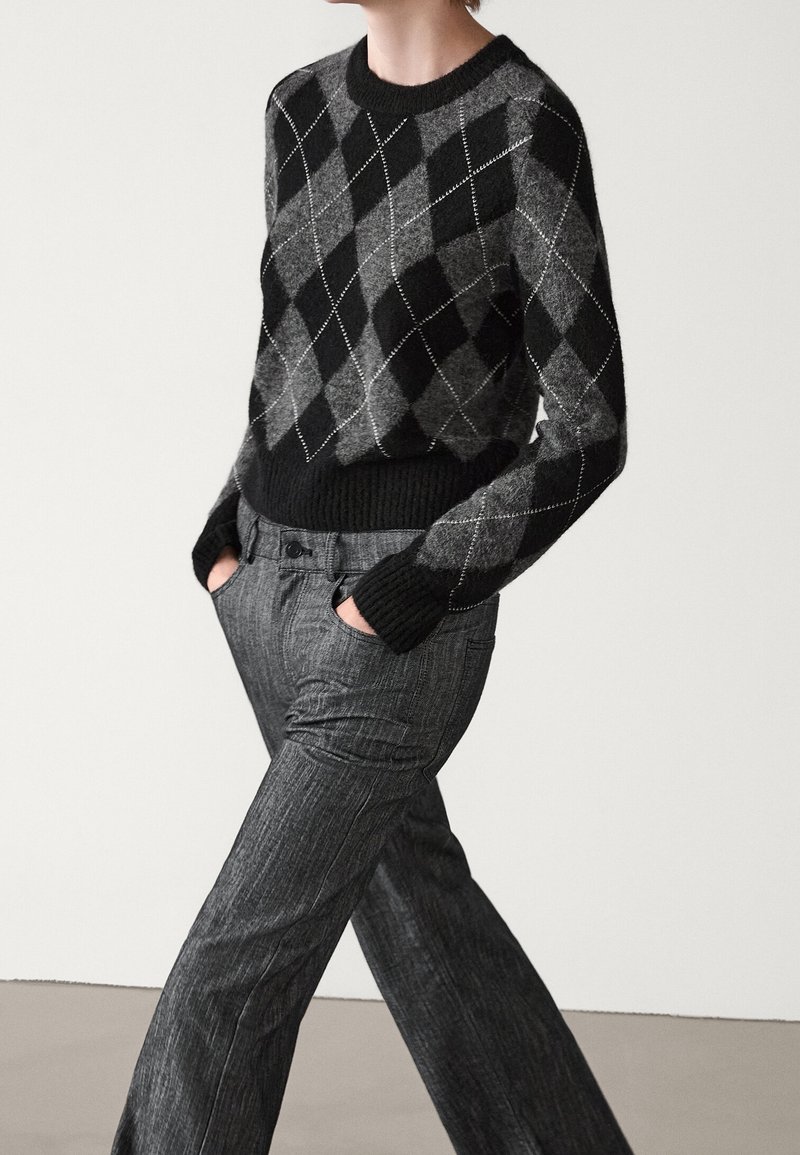 Personne portant un pull à losanges noir et gris avec un pantalon texturé gris, debout les mains dans les poches contre un fond uni.