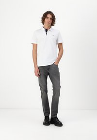 Camisa polo blanca con cuello azul marino, combinada con jeans gris oscuro. El modelo lleva zapatos negros y está de pie sobre un fondo claro y liso.
