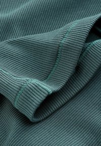 Žebrovaná tkanina v tmavě zelené barvě, s hladkým povrchem a kontrastním prošitím. Skládaný detail zdůrazňuje elasticitu materiálu.