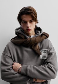 Grå sweatshirt med stor huva och en framstående etikett, med en brun stickad scarf hängande runt halsen. Slät textur, avslappnad passform.