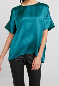 Top en satin turquoise avec une coupe décontractée, un col arrondi et des manches courtes, associé à un pantalon noir ajusté pour le contraste.
