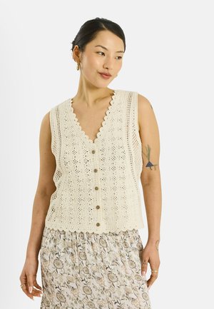 Femme aux cheveux courts et foncés portant un gilet crème en crochet boutonné et une jupe fleurie, debout devant un fond blanc.