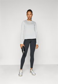 Világosszürke, hosszú ujjú felső, fekete leggings, egy fekete vízpalackot tart. Sportcipő szürke és fehér akcentusokkal. Simább anyag, testhezálló fazon.