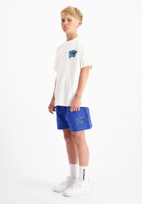 Witte t-shirt met blauwe graphic logo, gecombineerd met blauwe shorts met hetzelfde logo. Draagt witte sneakers en gestreepte sokken.
