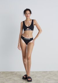 Crni bikini set s sportskim dizajnom, s braletom i donjim dijelom niskog struka. Tkanina izgleda glatko, s brendom u kontrastnoj bijeloj boji.