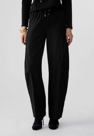 Donna che indossa pantaloni neri a gamba larga con vita con coulisse, scarpe nere con tacchi alti e braccialetti dorati al polso sinistro su sfondo semplice.