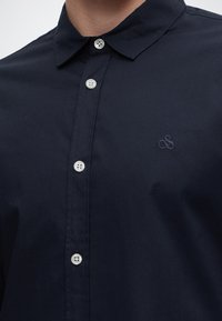 Gros plan d'une personne portant une chemise bleu marine foncé avec des boutons blancs et un petit logo brodé sur la poitrine.
