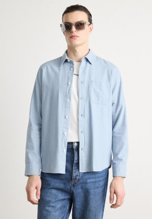 Light blue button-up shirt made of soft fabric, featuring a left chest pocket, paired with a white t-shirt and denim jeans.