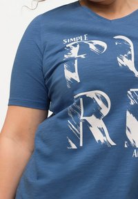 Blå kortärmad t-shirt med v-ringning, med stora vita bokstäver i en sliten stil. Tyget verkar mjukt och texturerat.