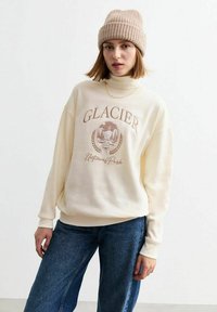 Sudadera color crema con cuello alto, que presenta un gráfico de "Parque Nacional GLACIER" en marrón. Combinada con jeans de pierna ancha azules y un gorro de punto beige.