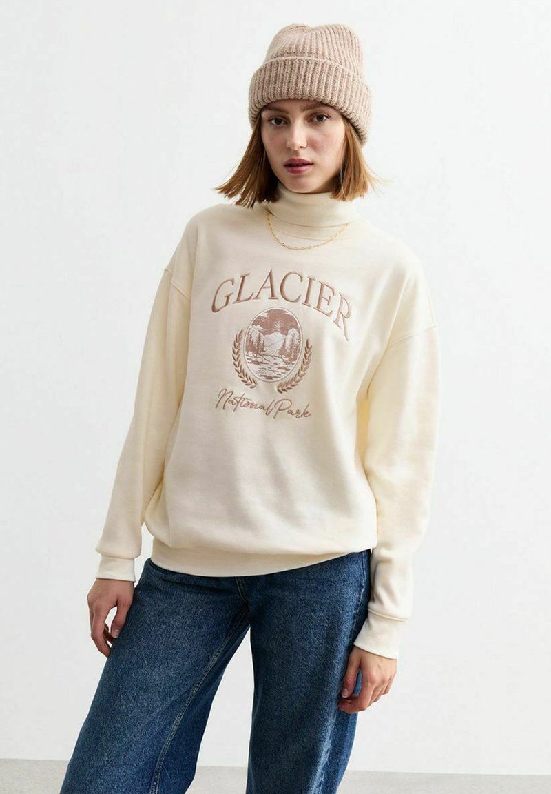 Sudadera color crema con cuello alto, que presenta un gráfico de "Parque Nacional GLACIER" en marrón. Combinada con jeans de pierna ancha azules y un gorro de punto beige.