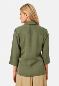 Olivgrünes, leichtes Hemd mit Kragen und drei Viertel Ärmeln. Verfügt über eine glatte Textur und einen abgerundeten Saum, keine sichtbaren Beschläge.