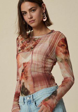 Un jeune mannequin pose dans un haut à manches longues à motif floral et un jean taille haute bleu clair, avec des cheveux légèrement coiffés et de grandes boucles d'oreilles.