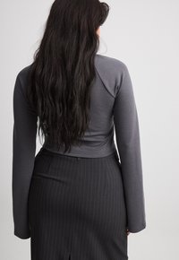 Frau mit langen dunklen Haaren, die ein eng anliegendes graues Langarmoberteil und einen hochgeschnittenen dunklen Nadelstreifenrock trägt, von hinten gezeigt.