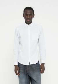 Νεαρός άνδρας που φοράει λευκό πουκάμισο με ρίγες και ξεβαμμένα μπλε τζιν, στέκεται μπροστά σε ένα απλό λευκό φόντο.