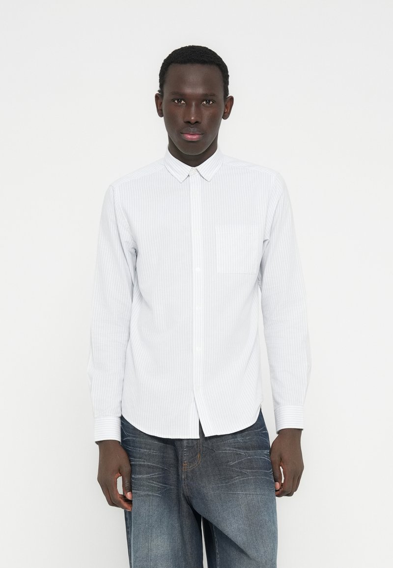 Νεαρός άνδρας που φοράει λευκό πουκάμισο με ρίγες και ξεβαμμένα μπλε τζιν, στέκεται μπροστά σε ένα απλό λευκό φόντο.