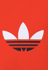 Röd tyg har en stor vit och svart Adidas trefoil-logotyp, med tre blad ovanför tre horisontella svarta ränder.