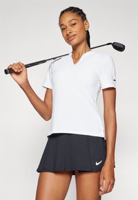 Balta trumpomis rankovėmis polo marškinėliai su tekstūruotais taškeliais, V formos iškirptė ir šoninės skeltukus; derinama su juoda sijonu, ant kurio matomas Nike logotipas. Laikoma golfo lazda.