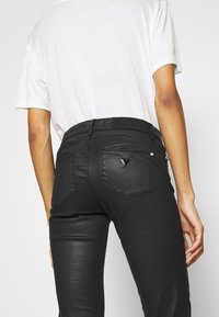 Jeans skinnies negros con acabado parecido al cuero, con bolsillos traseros y un pequeño logo en la cinturilla. Diseño simple y ajustado.