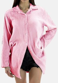 Lichtroze overhemd met knopen, gemaakt van een textuurrijke, gekreukte stof en lange mouwen. Heeft een puntkraag en zijslitten. Draagt boven een zwarte korte broek.