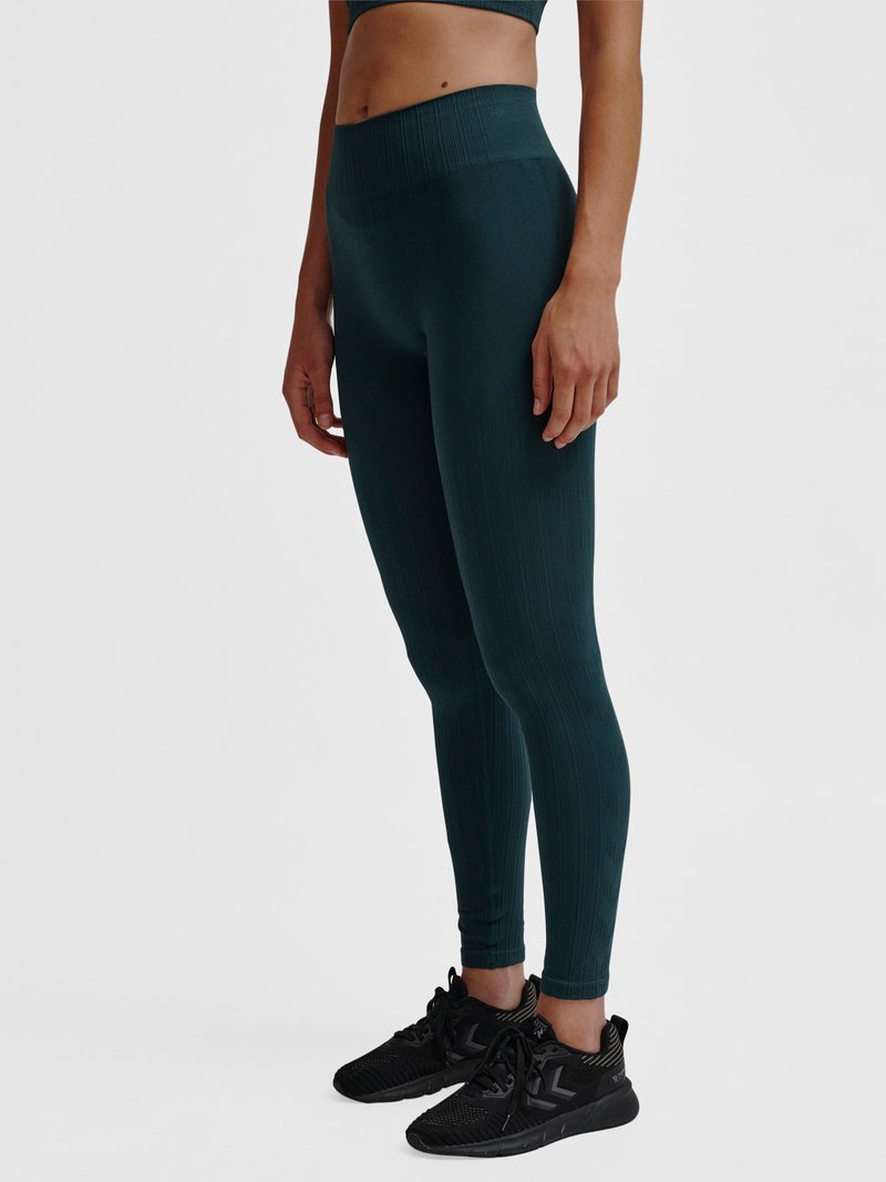 Kobieta modelka nosząca ciemnozielone teksturowane legginsy z wysokim stanem oraz czarne buty sportowe, stojąca na jednolitym jasnym tle.