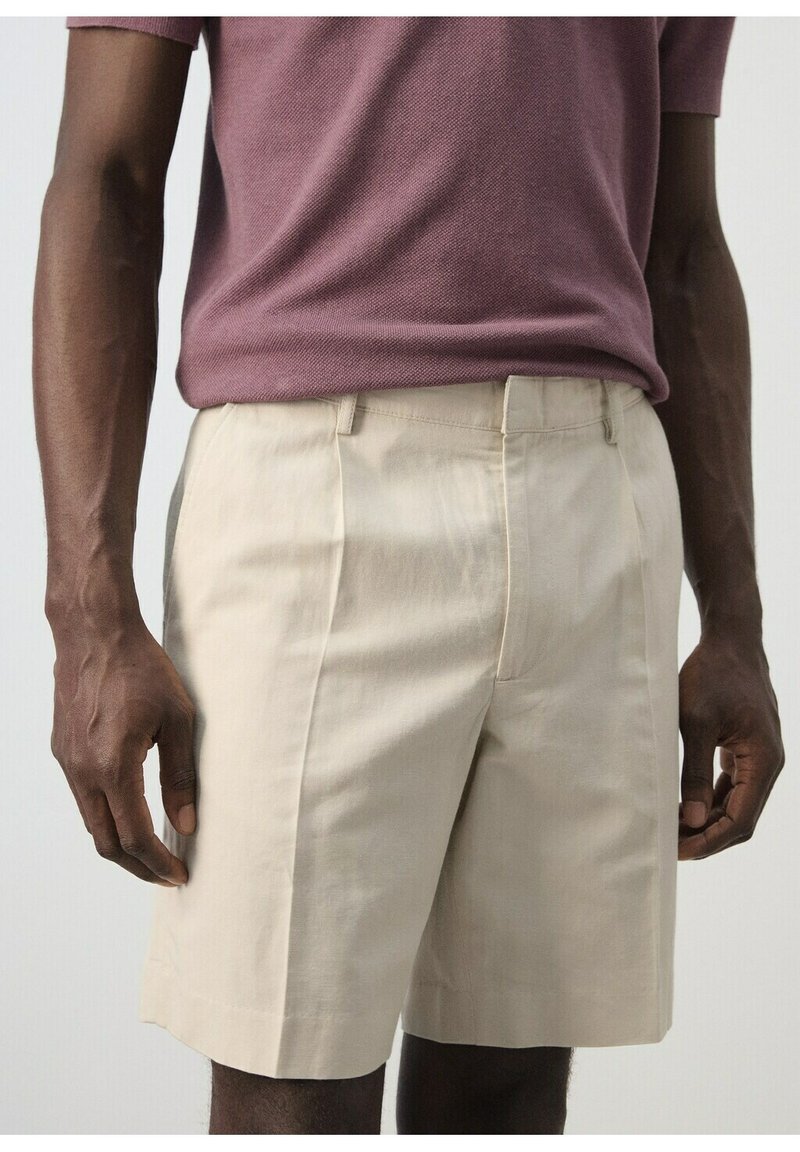 Homme portant un short beige sur mesure arrivant aux genoux et un pull à manches courtes mauve, debout les bras détendus le long du corps.